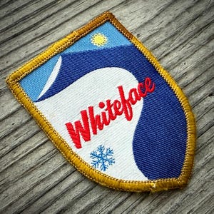 May include: Embroidered patch with a shield shape, featuring the word "Whiteface" in red script. The design includes a blue and white background with a sun and snowflake, framed by a gold border.
