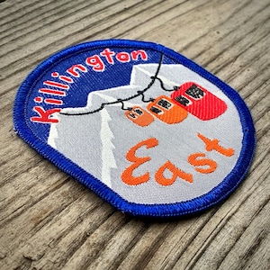 May include: A blue and white embroidered patch with the text "Killington East" and a depiction of a ski lift with three gondolas against a mountain backdrop.