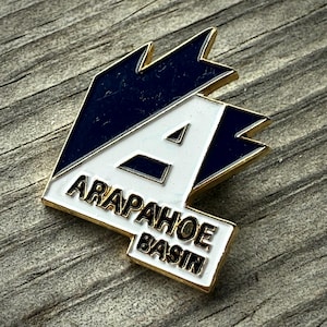 May include: A gold-edged pin featuring the text "ARAPAHOE BASIN" in black lettering. The pin has a white background with a blue and gold design. The letter "A" is prominently displayed in white.