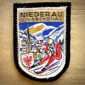 May include: A vintage patch with a black border features a ski scene. The patch includes the text "NIEDERAU WILDSCHONAU" and "TIROL" with a red eagle emblem. A skier in red skis down a snowy mountain.