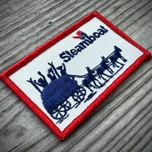 May include: A rectangular embroidered patch with a white background and a red stitched border. The design features dark blue embroidery depicting a horse-drawn wagon and the text "Steamboat" with a small red accent above the letter S.