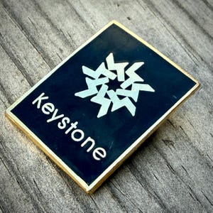 May include: A rectangular pin with a dark blue background and a gold border. The pin features the word "keystone" in white, along with a white geometric design. The pin is set on a wooden surface.