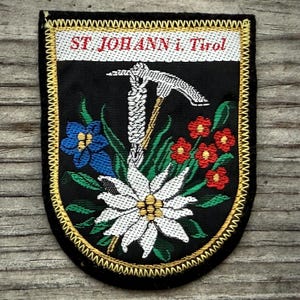 May include: Embroidered patch with a black background, featuring the text "ST JOHANN i. Tirol" in white. The patch includes a white edelweiss flower, blue and red flowers, and a pickaxe design. The border is gold.