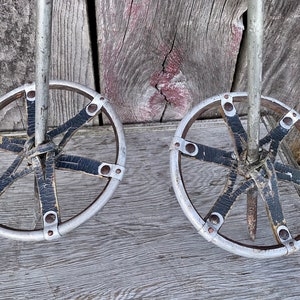 May include: Pair of vintage ski poles with circular baskets. The poles have silver shafts and circular baskets with black leather straps. The background is weathered wood.