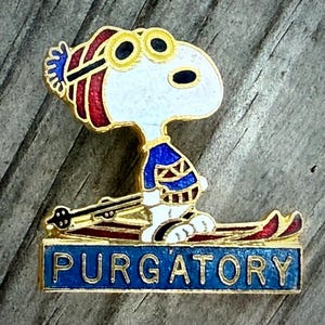 May include: A Snoopy pin featuring Snoopy in a blue and gold ski outfit with goggles, skis, and the word "PURGATORY" on a blue banner. The pin has gold trim and red accents.