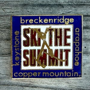 May include: A rectangular pin with a blue border and white center. The pin features the words "Breckenridge," "Ski the Summit," and "Copper Mountain." A skier graphic is in gold. The pin also includes the words "Keystone" and "Arapahoe."