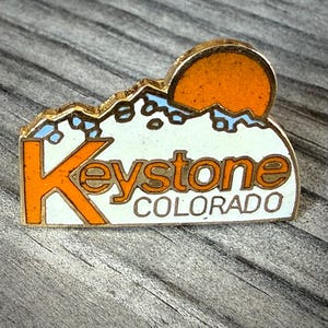 May include: A vintage Keystone Colorado souvenir pin. The pin features an orange sun setting behind white mountains with blue accents. The word "Keystone" is in orange, and "Colorado" is in white.