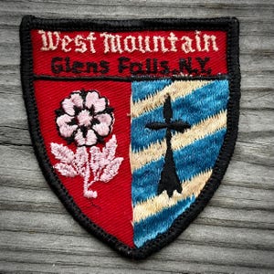 May include: A shield-shaped embroidered patch with a black border. The top red section features white text reading West Mountain Glens Falls, N.Y. The bottom is split into a red side with a pink flower and a blue and cream striped side with a black cross.