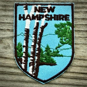 May include: A shield-shaped embroidered patch with a light blue background featuring white and brown birch trees, green pine trees, and the text NEW HAMPSHIRE in dark brown letters. The patch has a dark brown stitched border.