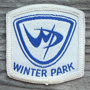 May include: A square embroidered patch with rounded corners featuring a white background and a blue border. The center displays a blue shield shape containing a stylized blue WP logo. The text WINTER PARK is embroidered in blue below the shield.