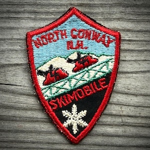 May include: A shield-shaped embroidered patch with a red border. The top features light blue sky with the text NORTH CONWAY N.H. in red. Below are white snowy mountains, two red ski cars, a green lattice structure, and a white snowflake on black.