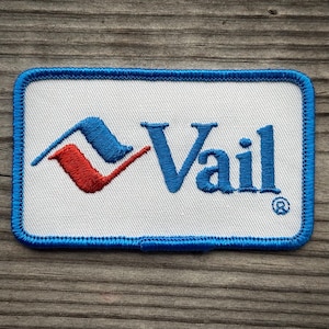 May include: A rectangular embroidered patch with a white background and a blue stitched border. It features a blue and red mountain-shaped logo to the left of the blue embroidered text Vail, followed by a small registered trademark symbol.