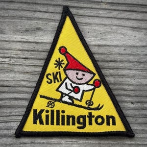 May include: Yellow triangular patch with a black border featuring a cartoon skier in a red hat and white outfit. The patch reads "SKI" and "Killington" in black text. The skier is on skis with poles.