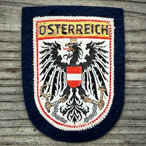 May include: A navy blue patch with the word "ÖSTERREICH" in gold lettering. The patch features the Austrian coat of arms, a black eagle with a red and white shield, and gold accents.