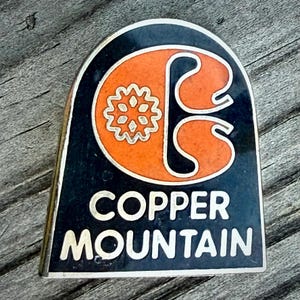 May include: A vintage Copper Mountain ski resort pin. The pin features a black background with the words "Copper Mountain" in silver lettering. An orange and silver graphic of a stylized "C" and a flower is in the center.