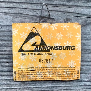 May include: A yellow ski ticket with a black mountain and ski slope graphic. The text "ANNONSBURG" is printed in black letters. The text "SKI AREA AND SHOP" is printed in black letters. The ticket number "087617" is printed in black letters. The text "The purchaser and user of this ticket assumes and understands that skiing is a has sport, and that bare spots, variations in snow, ice and terrain along with bumps, moguls, stumps, forest growth and debris and rocks and many other natural or manmade hazards or obstacles exist within this ski area. In using the ticket and skiing at the area, such dangers are recognized and accepted whether they" is printed in black letters. The text "VOID IF DI" is printed in black letters.