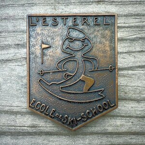 May include: A bronze-colored metal plaque with the words "L'ESTEREL ECOLE DE SKI-SCHOOL" in relief. The plaque features a skier graphic and a small flag. The plaque is mounted on a weathered wooden surface.