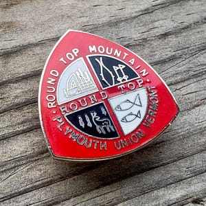 May include: A red, white, and black Round Top Mountain Plymouth Union Vermont pin. The pin is triangular with a gold border and features a segmented design with various illustrations and text.