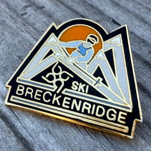May include: A vintage Breckenridge Ski pin with a black border and gold accents. The pin features a skier, mountains, and the words "SKI BRECKENRIDGE". The background is orange and the skier is wearing blue. The pin is triangular in shape.
