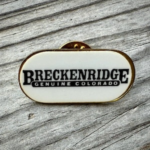 May include: A rectangular pin with a white background and gold trim. The pin features the text "BRECKENRIDGE GENUINE COLORADO" in black, bold letters. The pin is set against a weathered wooden surface.