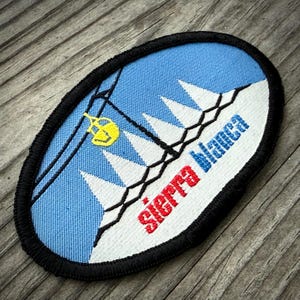 May include: Oval patch with a blue background featuring a ski lift and mountain peaks. The patch has the words "Sierra Blanca" in red and blue. The patch is outlined in black.