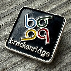 May include: A square, black enamel pin with a silver border. The pin features the word "breckenridge" in silver lettering. The letters "bo99" are in blue, green, yellow, and red.