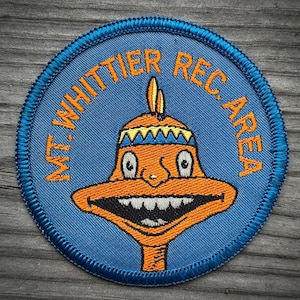 May include: A circular embroidered patch with a blue background and a dark blue stitched border. The patch features an orange cartoon character with a wide grin, white teeth, and a yellow and blue headband. The text MT. WHITTIER REC. AREA is in orange.