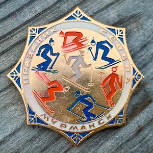 May include: A gold-colored metal pin with a white circle in the center. The circle features five skiers in red, blue, and gray, with the text "ПРАЗДНИК С СВЕЕРДА" around the outside. The text "МУРМАНСК" is at the bottom of the circle.