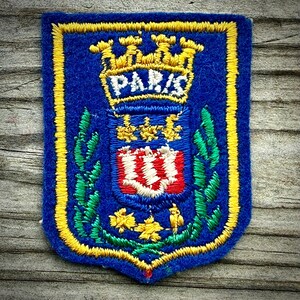 May include: A rectangular blue embroidered patch featuring a yellow border and a central crest. The crest includes a yellow crown above the text PARIS, a red and white shield, green leafy branches on the sides, and yellow floral accents below.