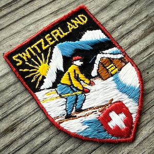 May include: Embroidered patch featuring a skier on a snowy mountain scene with the word "SWITZERLAND" in yellow. The patch has a red border and includes a Swiss flag. The design incorporates blue, white, yellow, and black.