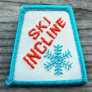 May include: Embroidered patch with a white background, a turquoise border, and the words "SKI INCLINE" in orange. A turquoise snowflake is embroidered below the text. The patch is rectangular with a trapezoid shape.