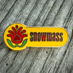 May include: A yellow and red Snowmass pin. The pin features a red flower design with green accents on the left side and the word "Snowmass" in red letters on the right. The pin is set against a weathered wood surface.