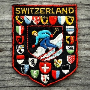 May include: Embroidered patch with the word "SWITZERLAND" in gold lettering. The patch features a skier in blue and a collection of colorful shield emblems. The patch has a black background with a red border.
