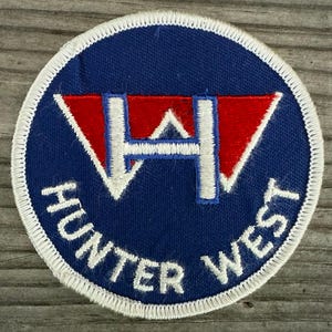May include: A circular patch with a blue background and white stitched border. The patch features a red and white design with the letters "H" and "W" and the words "HUNTER WEST" in white lettering.