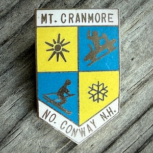 May include: A vintage enamel pin with a shield design. The pin features the text "MT. CRANMORE" at the top and "NO. CONWAY N.H." at the bottom. The shield is divided into four quadrants with yellow and blue backgrounds, and ski-themed graphics.