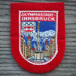 May include: A red felt patch with a white stitched border. The patch features a detailed embroidered image of a city scene with mountains, buildings, and the Olympic rings, with the text "OLYMPIASTADT INNSBRUCK 1975".