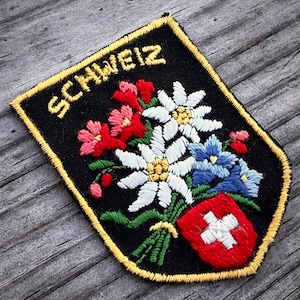 May include: Embroidered patch with the word "SCHWEIZ" in gold lettering. The patch features a bouquet of red, white, and blue flowers, a red shield with a white cross, and a gold border on a black background.