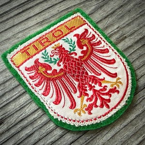 May include: Embroidered patch featuring a red eagle emblem with gold accents, the word "TIROL" in gold, and a green border. The patch is white with red stitching and a green felt edge.