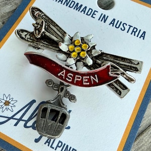 May include: A silver-toned metal pin featuring skis, an edelweiss flower, and a red banner with "ASPEN" in white lettering. A small gondola charm hangs below. The pin is on a card that says "HANDMADE IN AUSTRIA".
