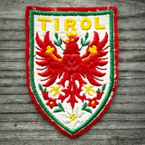May include: Embroidered patch featuring a red eagle with yellow accents, the word "TIROL" in yellow, and a green and red border. The patch has a white background and is shaped like a shield.