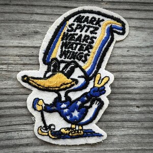 May include: Embroidered patch featuring a cartoon duck wearing water wings. The patch has a white background with black, blue, and yellow accents. Text on the patch reads "MARK SPITZ WEARS WATER WINGS". The duck is holding up a peace sign.