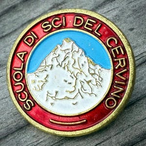 May include: A round pin with a red and gold border, featuring the text "SCUOLA DI SCI DEL CERVINO." The center depicts a mountain scene with a blue sky and white snow-capped peaks. The pin has a gold-colored edge.