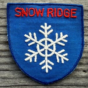 May include: A blue patch with the red text "SNOW RIDGE" above a white snowflake design. The patch has a shield shape and is likely an emblem or badge.