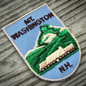 May include: A shield-shaped embroidered patch with a light blue background. It features a green mountain graphic with white highlights and a brown train track. The text reads MT. WASHINGTON in brown at the top and N.H. in brown at the bottom.