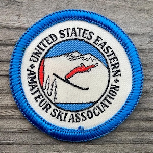 May include: A white and blue embroidered patch with the text "United States Eastern Amateur Ski Association". The patch features a skier in red and black, skiing down a mountain.