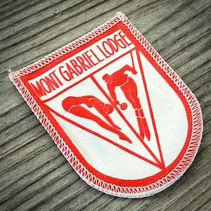 May include: A white and red patch with the text "MONT GABRIEL LODGE" and illustrations of divers. The patch has a red border with white stitching and is set against a wooden background.
