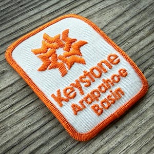 May include: Embroidered patch with a white background and orange border. The patch features orange starburst designs and the words "Keystone Arapahoe Basin" in orange lettering. The patch is rectangular with rounded corners.