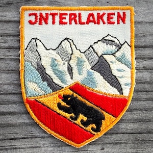 May include: Embroidered patch with the word "INTERLAKEN" in red. The patch features a mountain range in shades of gray and blue above a shield with a black bear on a red and yellow background. The patch has a yellow border.