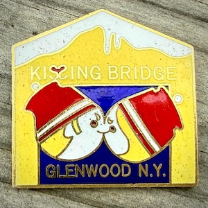 May include: A rectangular pin with a yellow background and white trim. The pin features the words "Kissing Bridge" and "Glenwood N.Y." in gold lettering. Two cartoon figures are kissing. The figures are wearing red hats and are kissing.