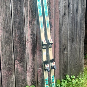 May include: A pair of vintage wooden skis with blue and white graphics. The skis have black bindings and the text "TR COMP" is printed on the side.